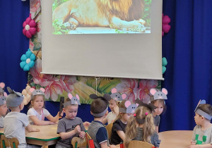 Dzieci siedzą przy stolikach, w tle lew.