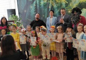 Zdjęcie przedstawia wszystkich uczestników konkursu wokalnego stojących na scenie z dyplomami w dłoniach, wraz z jury oceniającym zmagania dzieci.