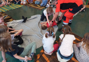 Ratownik medyczny instruuje dzieci jak ułożyć osobę poszkodowaną w pozycji bocznej ustalonej.