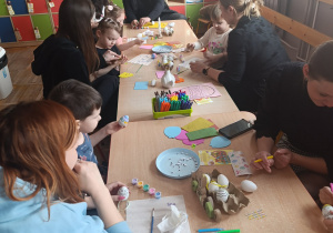 Rodzice siedzą wspólnie z dziećmi przy stole i ozdabiają pisanki.