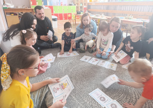 Dzieci i rodzice siedzą na dywanie i wspólnie grają z rodzicami.