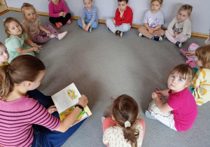 Dzieci siedzą w kole i słuchają bajki czytanej przez starszą dziewczynkę.