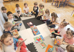 Dzieci siedzą w kole na dywanie i oglądają przyniesione książki z bajkami.