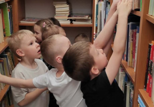 Dzieci przyglądają się półkom z książkami w szkolnej bibliotece, jeden chłopiec wyjmuje z półki książkę.