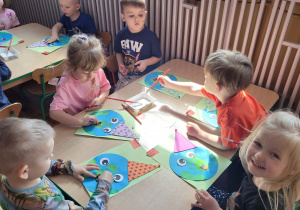 Dziewczynki i chłopcy siedzą przy stoliku i wykonują pracę plastyczną.