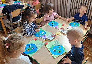 Dziewczynki i chłopcy siedzą przy stoliku i wykonują pracę plastyczną.