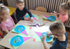 Dziewczynki i chłopcy siedzą przy stoliku i wykonują pracę plastyczną.