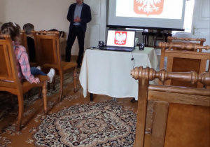 Pracownik muzeum stojący obok ekranu projekcyjnego, na którym wyświetlany jest herb Polski.
