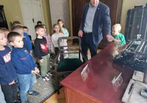 Dzieci oglądają dawna maszynę do pisania będącą na wyposażeniu muzeum oraz słuchają informacji na jej temat.