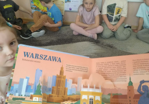 Zdjęcie przedstawia dzieci siedzące na dywanie a przed nimi stoi książka Polska w stylu pop -up i wykonują zadania jakie przed nimi stawia projekt