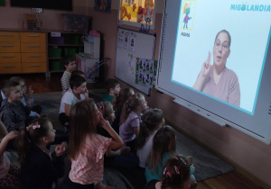 Na zdjęciu widzimy dzieci siedzące przed tablicą multimedialną i oglądające jak pani pokazuje w języku migowym słowo mama