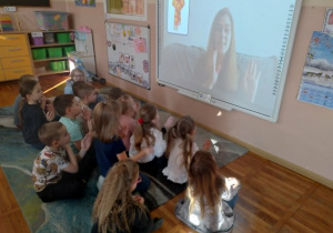 Na zdjęciu widzimy dzieci siedzące na dywanie przed tablicą multimedialną które patrzą jak w języku migowym pokazać słowo szalik