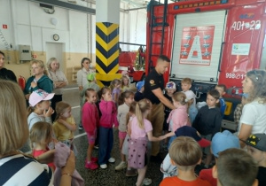 Na zdjęciu widzimy strażaka stojącego przed wozem strażackim który wyjaśnia dzieciom co się znajduje we wnętrz samochodu pożarniczego