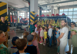 Zdjęcie przedstawia dzieci stojące w remizie strażackiej które zapoznają się z ubiorem strażaka