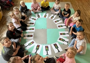 Dzieci z grupy Żabki pozują do zdjęcia siedząc na dywanie z sygnalizatorami świetlnymi.