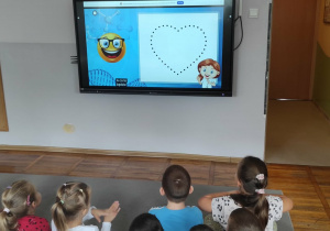 Dzieci z grupy Misie siedzą na dywanie oglądając film edukacyjny wyświetlany na tablicy multimedialnej dotyczący Dnia Kropki.