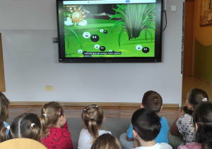 Dzieci z grupy Misie siedzą na dywanie oglądając film edukacyjny wyświetlany na tablicy multimedialnej dotyczący Dnia Kropki.