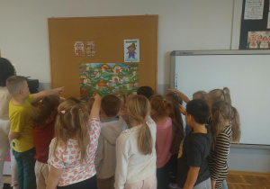 Na zdjęciu widzimy dzieci stojące przodem do tablicy na której zawieszony jest plakat z wioską Kumpelków. Dzieci wskazują palcami na plakat