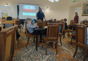 Na zdjęciu widzimy dzieci siedzące na krzesełkach w sali muzeum przed nimi stoi Pan który prowadzi warsztaty i wyświetla prezentacje na ekranie.