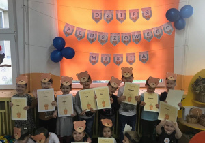 Dzieci maja na głowach opaski z głową brązowego misia, w rękach trzymają dyplomy. Na rolecie widac napis Dzień Pluszowego Misia.