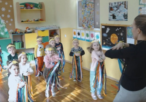 Dzieci i nauczycielka z grupy Słoneczka trzymaja zaplecione przed sobą ręce i tańczą z kolorowymi wstążkami.