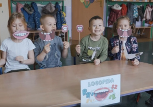 Na zajęciach logopedycznych dzieci siedzą przy stoliku i przykładają do twarzy wycięte uśmiechyi inne minki symbolizujące ćwiczenia buzi i języka..