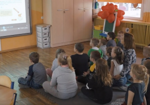 Dzieci siedza na macie i patrzą na tablicę multimedialną, na której przedstawiono treści edukacyjne o dinozaurach.