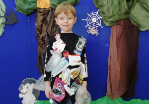 Chłopiec grający Eko – Smyka w kamizelce, do której przyszyte są różnego rodzaju śmieci stojący na tle dekoracji przedstawiającej las.