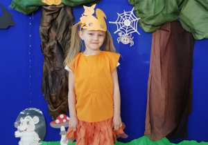 Dziewczynka grająca drugą wiewiórkę ubrana na pomarańczowo i w masce wiewiórki, stojąca na tle dekoracji przedstawiającej las.