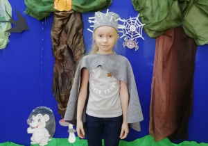 Dziewczynka przebrana za jeża ubrana w szarą pelerynkę oraz opaską na głowie z kolcami z plastikowych słomek stojąca na tle dekoracji lasu.