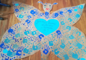 Plakat wykonany przez grupę Smerfy. Wycięty z szarego papieru motyl wypełniony odbitymi na niebiesko dłońmi dzieci a w środku przyklejone niebieskie serduszko z napisem: „Jesteśmy z Wami niebieskimi Motylami”.