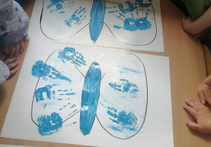 Prace plastyczne w wykonaniu grupy Biedronki. Trzy plakaty z konturami motylka wypełnionymi odciśniętymi rączkami dzieci. Na górze pierwszego kartonu napis : Jesteśmy z Wami Niebieskimi Motylami.