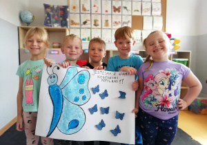 Piątka dzieci stoi w niebieskich koszulkach i trzyma wykonany plakat z niebieskim motylem i małymi motylkami oraz napisem „Jesteśmy z Wami niebieskimi motylami”.