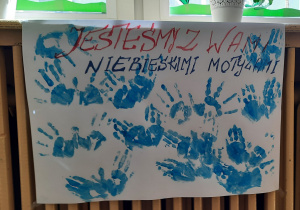 Plakat wykonany przez grupę Misie. Na białym kartonie odbite na niebiesko dłonie dzieci oraz na górze napis czerwono – niebieski : „Jesteśmy z Wami Niebieskimi Motylami”.