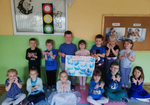 Siedmioro dzieci stoi pod ścianą a sześcioro siedzi na matach ubranie w niebieskie koszuli. Trójka dzieci trzyma wykonany plakat a reszka pokazuje dłonie ułożone w kształcie motylka.