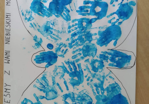 Plakat wykonany przez grupę Motylki. Narysowany kontur motyla na białym kartonie wypełniony odbitymi na niebiesko dłońmi dzieci a na górze napis: „Jesteśmy z Wami niebieskimi Motylami”.
