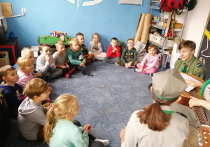Dzieci i harcerze siedzą w kręgu w harcówce na dywanie.