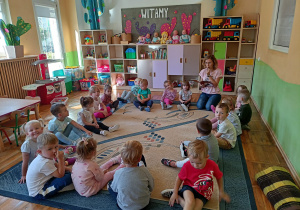 Przedszkolaki siedzą na dywanie isłuchaja jak nauczycielka czyta bajkę.