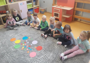 Dzieci siedzą na dywanie, do bluzek mają dopięte koła z literami tworzącymi napis „Dzień Kropki”.