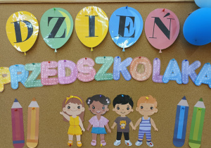 Na tablicy znajduje się przygotowana dekoracja tematyczna z okazji Dnia Przedszkolaka – Napis oraz uśmiechnięte sylwety dzieci i kolorowe kredki z papieru.
