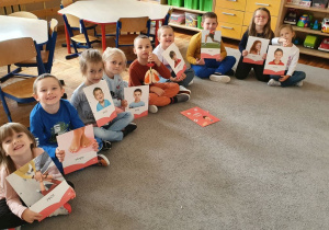 Grupa dzieci siedzi na dywanie. W dłoniach dzieci trzymają karty, na których są ilustracje przedstawiające części ciała.