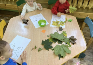 Dzieci wykonują pracę plastyczną – przyklejają suche liście na kartce z sylwetą twarzy dziecka.