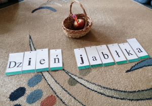 Na dywanie stoi pleciony koszyczek z jabłkiem i robaczkiem z drucika kreatywnego. Pod spodem napis ułozony z literek - Dzień jabłka.