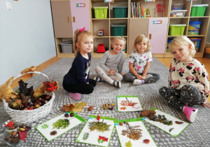 Dzieci siedzące wśród ilustracji drew i jesiennych darów.
