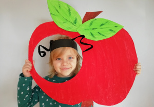 Dziewczynka w opasce z czułkami trzyma głowę w konturze czerwonego papierowego jabłka.