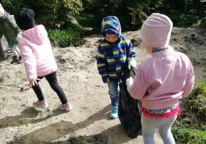 Chłopiec trzyma foliowy worek, a dzieci dookoła poszukują śmieci.