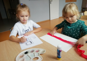 Dwie dziewczynki siedzą przy stoliku jedna przykleja czerwoną bibułę do kartki, a druga czarną farbą wykropkowuje kontu symbolu Polski Walczącej.