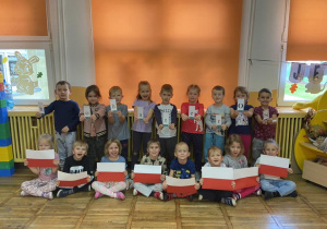 Grupa dzieci pozuje do zdjęcia. Część dzieci trzyma karteczki z literami, które układają się w napis „BohaterOn”, reszta dzieci trzyma flagę Polski.