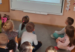 Dzieci siedzą na dywanie, oglądają projekcję filmu „Niebajka o Powstaniu Warszawskim”.