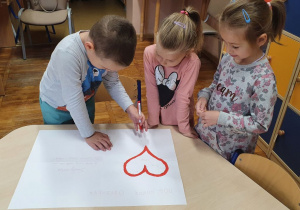 Dwie dziewczynki i chłopiec podpisują się na białej kartce z czerwonym sercem.